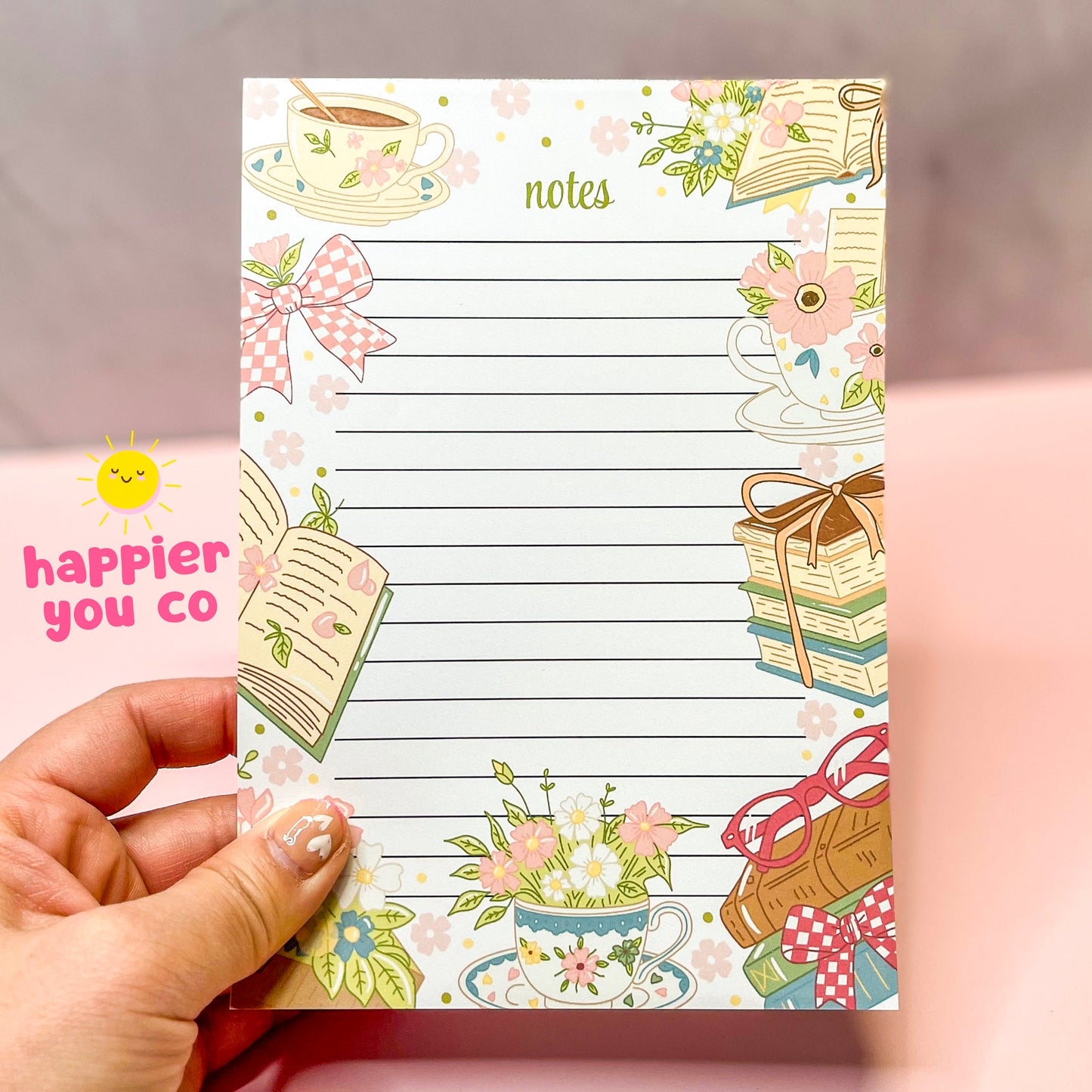 Decorative notepad with floral and book illustrations held by a hand, with text 'happier you co'