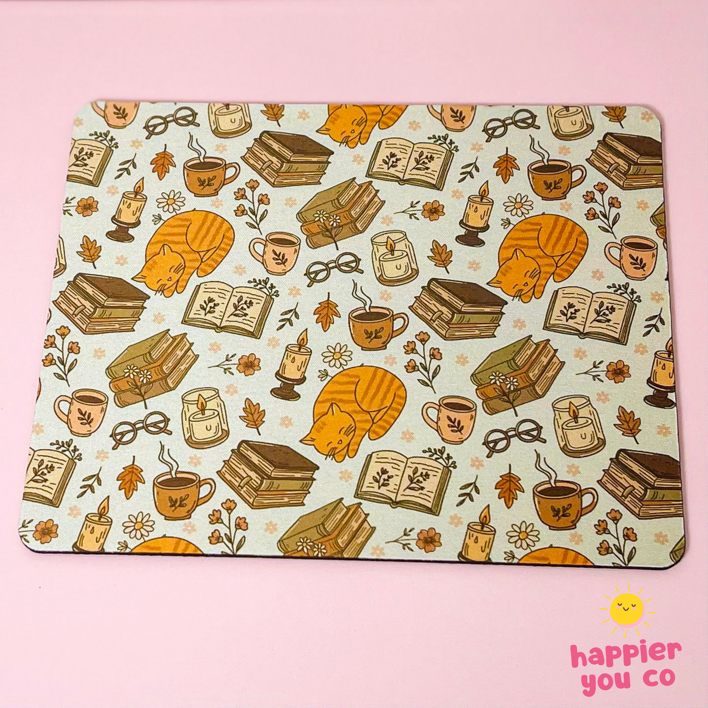 A rectangular desk mat with rounded corners featuring a cozy autumn-themed pattern of orange sleeping cats, open books, coffee cups, candles, and books on a light cream background. The desk mat is photographed flat on a solid pink background, with a 'happier you co' logo in the bottom right corner.
