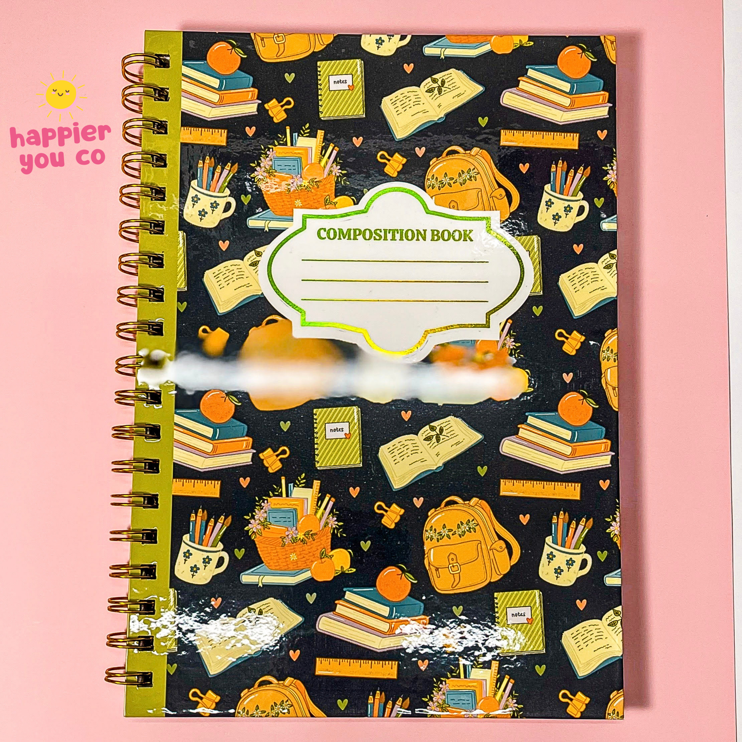 A spiral-bound composition notebook with a black cover decorated with a colorful school-themed pattern of books, backpacks, pencils, and cups. The notebook has a white label area in the center with 'COMPOSITION BOOK' printed on it.