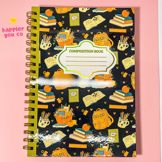 A spiral-bound composition notebook with a black cover decorated with a colorful school-themed pattern of books, backpacks, pencils, and cups. The notebook has a white label area in the center with 'COMPOSITION BOOK' printed on it.