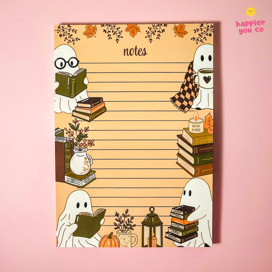 A rectangular notepad with a warm beige background and lined writing area, decorated with cute ghost illustrations, books, flowers, and autumn leaves. The word 'notes' appears at the top center.