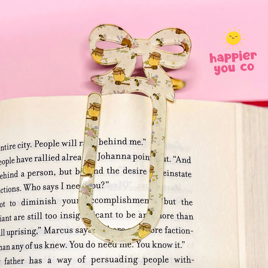 Decorative bookmark with floral design on an open book, against a pink background with 'happier you co' branding.