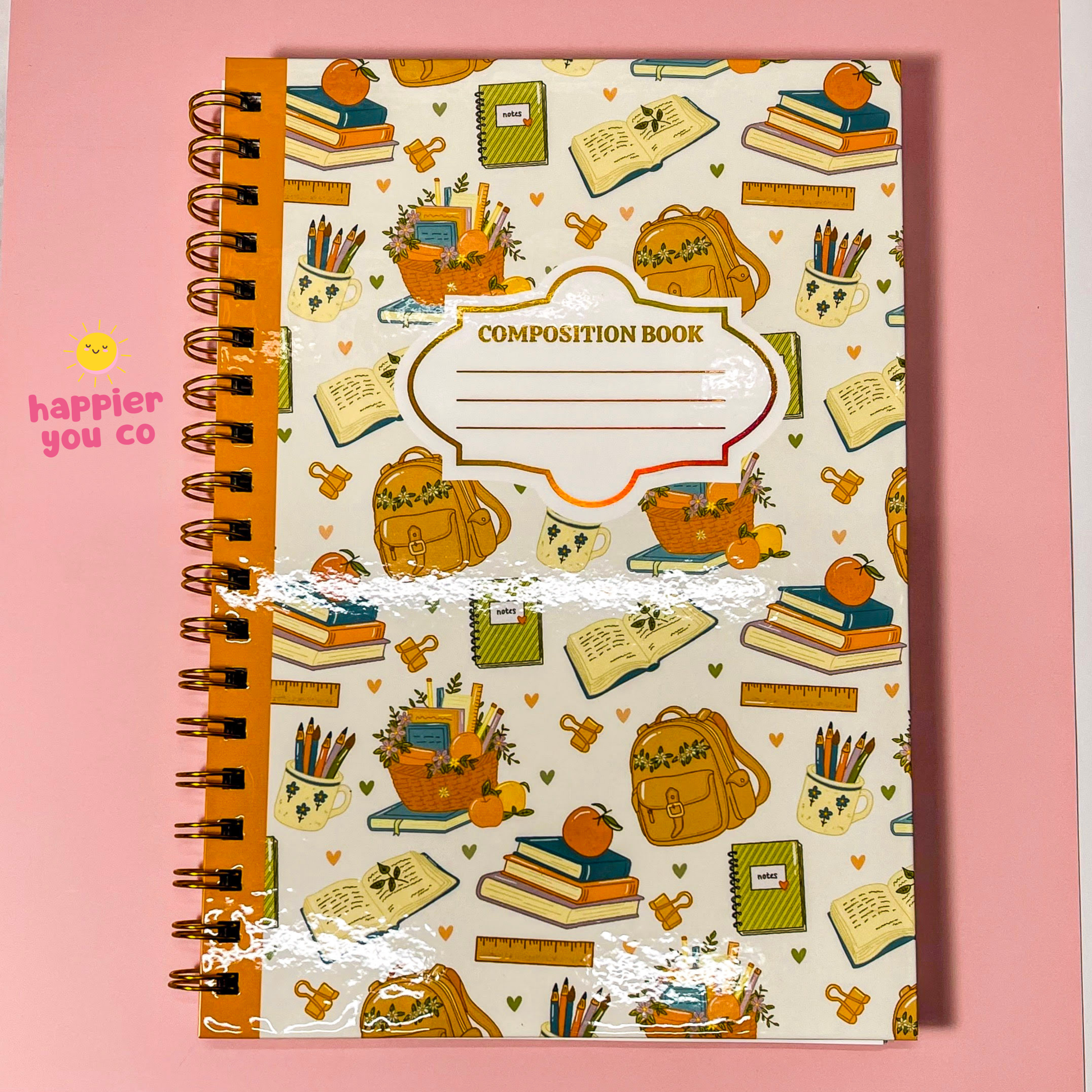 A spiral-bound composition notebook with a cream cover decorated with a colorful school-themed pattern of backpacks, books, pencils, and notebooks, set against a solid pink background.