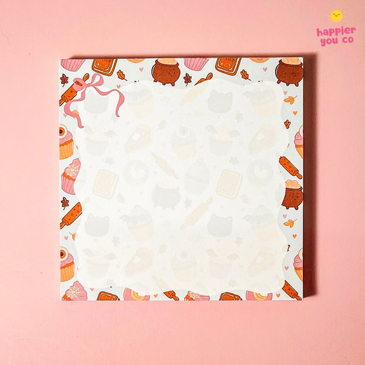 A square notepad with a cute pastel patterned border featuring cupcakes, cookies, and small illustrations, set against a solid pink background. The brand name 'happier you co' is visible in the top right corner.