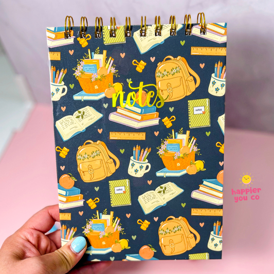 A hand with light blue nail polish holds a spiral-bound notebook featuring a navy blue background with a colorful school-themed pattern of backpacks, books, pencils, and flowers. The word 'notes' is written in yellow script in the center.