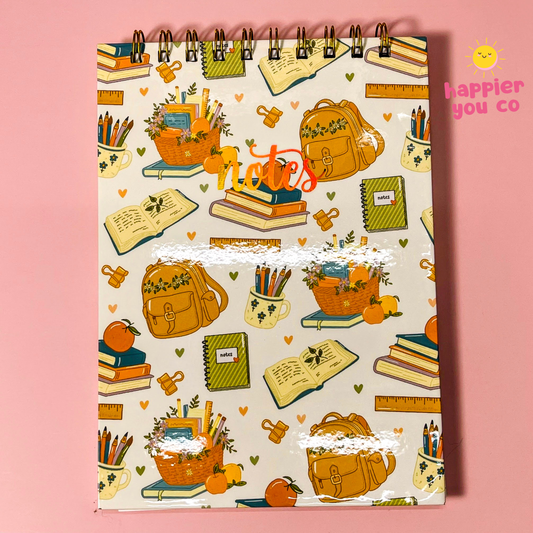 A spiral-bound notebook with a cream cover featuring a colorful school-themed pattern of backpacks, books, pencils, and notebooks, set against a solid pink background. A 'happier you co' logo is visible in the top right corner.