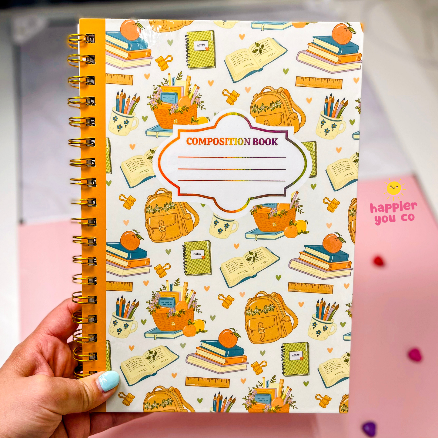 A hand with light blue nail polish holds a spiral-bound composition notebook. The notebook cover is white with a colorful pattern of school supplies like backpacks, books, pencils, and rulers, and features a central label that reads 'COMPOSITION BOOK'.