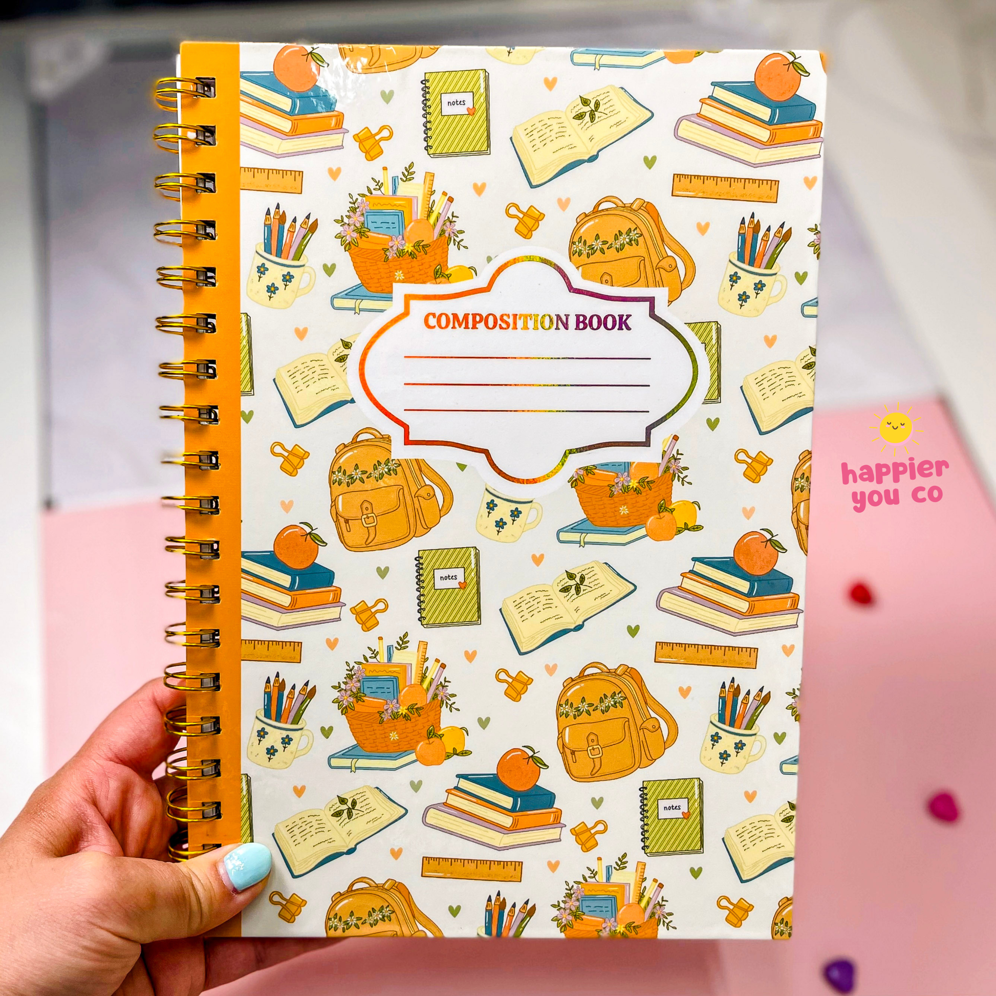 A hand with light blue nail polish holds a spiral-bound composition notebook. The notebook cover is white with a colorful pattern of school supplies like backpacks, books, pencils, and rulers, and features a central label that reads 'COMPOSITION BOOK'.