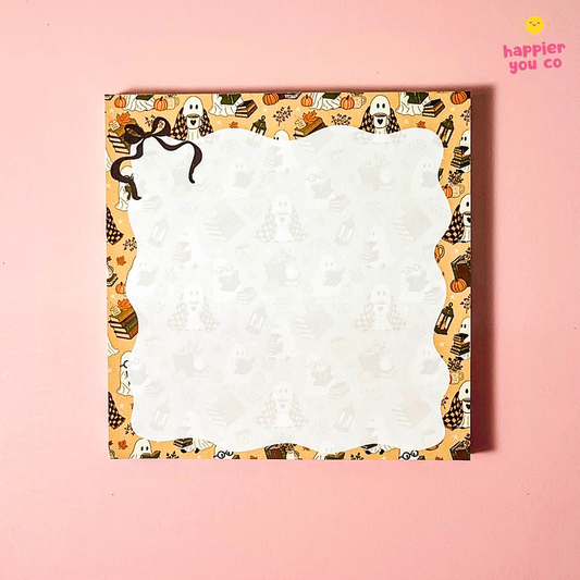 A square notepad with a scalloped white writing area and a Halloween-themed patterned border featuring ghosts, pumpkins, bats, and spiderwebs, shown on a solid pink background.