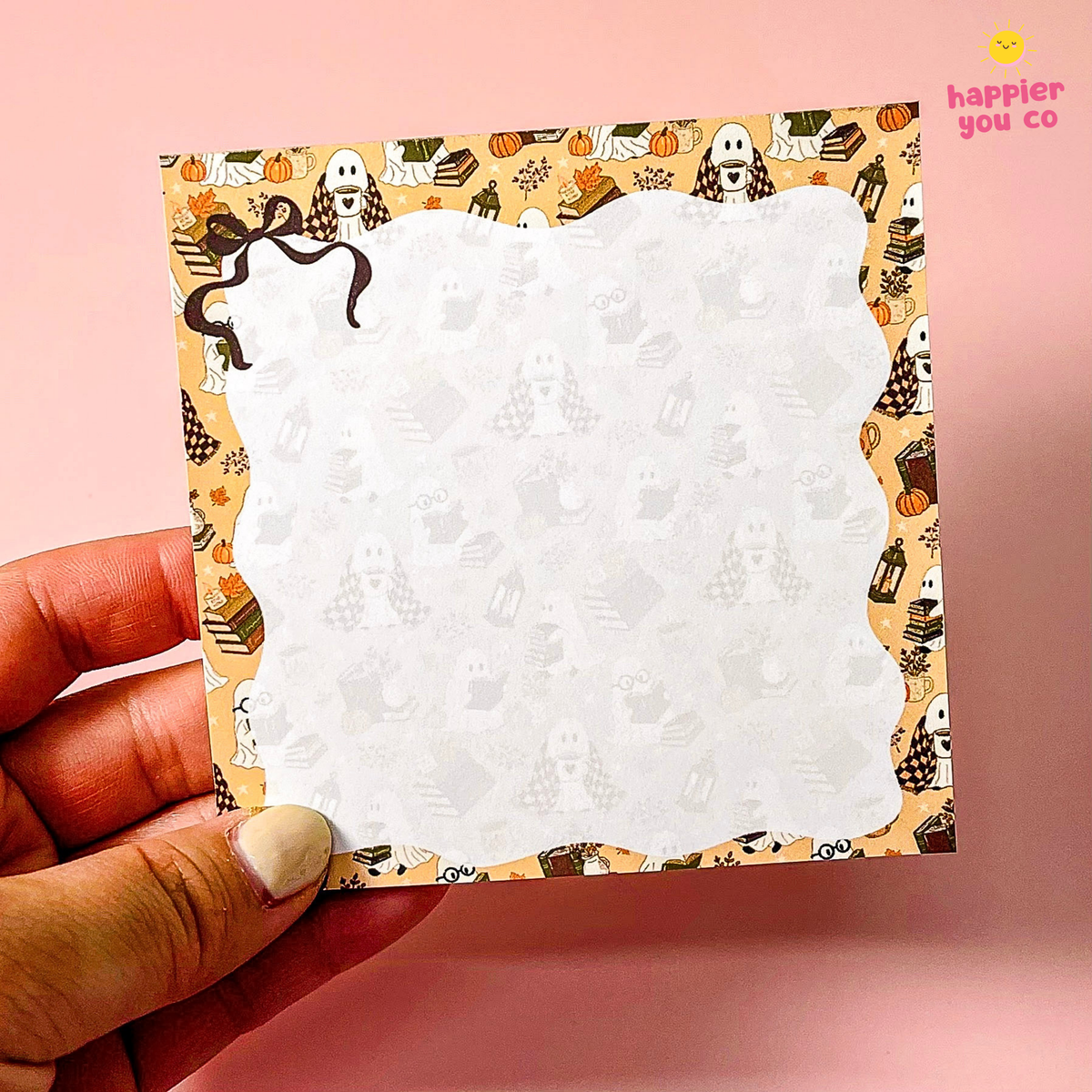 A hand holds a square notepad with a scalloped white writing area and a Halloween-themed patterned border featuring ghosts, pumpkins, and autumn motifs, set against a solid pink background.