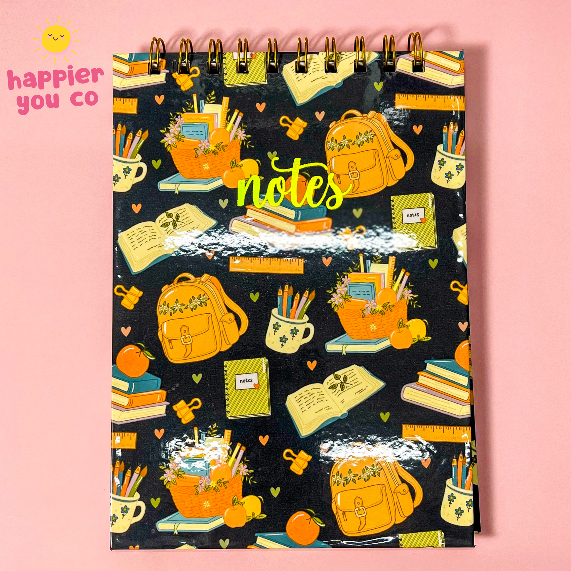 A spiral-bound notebook with a black cover decorated with a colorful school-themed pattern of backpacks, books, pencils, and flowers, set against a solid pink background. The brand logo 'happier you co' is visible in the top left corner.
