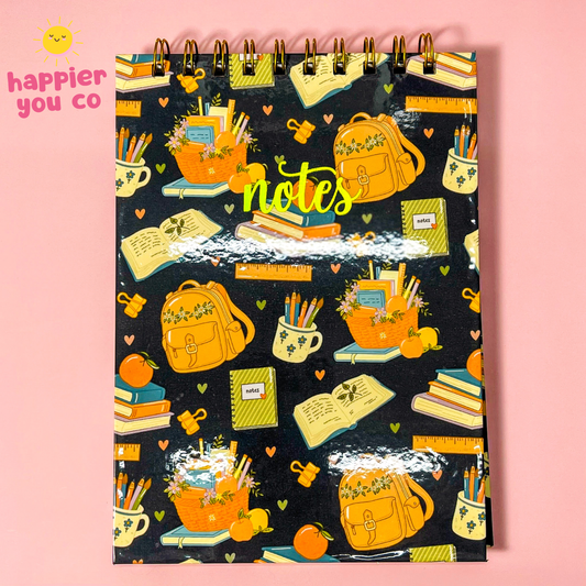 A spiral-bound notebook with a black cover decorated with a colorful school-themed pattern of backpacks, books, pencils, and flowers, set against a solid pink background. The brand logo 'happier you co' is visible in the top left corner.
