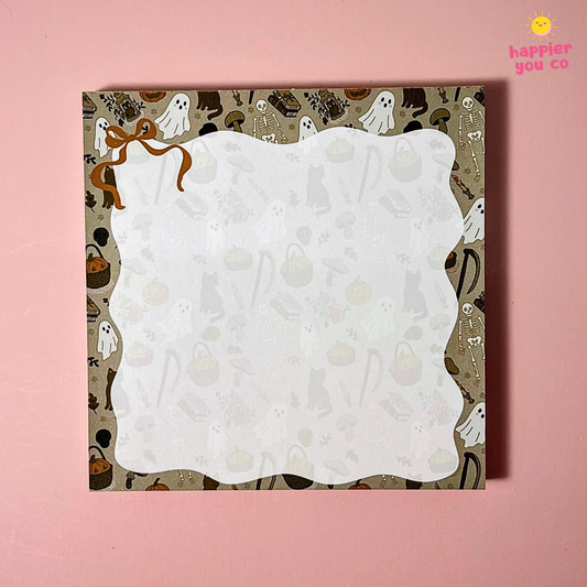 A square white card with scalloped edges and a Halloween-themed patterned border featuring ghosts, pumpkins, skeletons, and a brown ribbon illustration, displayed on a solid pink background.
