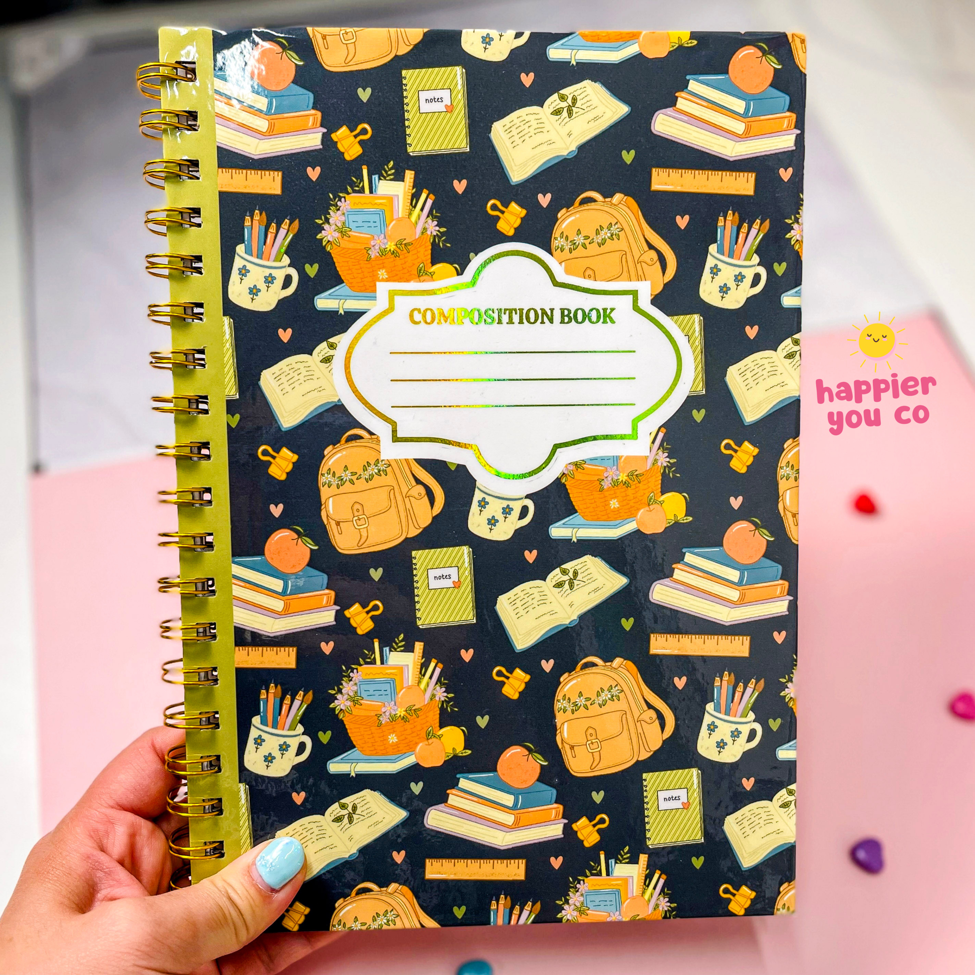 A hand with blue nail polish holds a spiral-bound composition book. The book cover is dark navy blue with a colorful pattern of school supplies like backpacks, books, pencils, and notebooks.