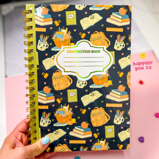 A hand with blue nail polish holds a spiral-bound composition book. The book cover is dark navy blue with a colorful pattern of school supplies like backpacks, books, pencils, and notebooks.