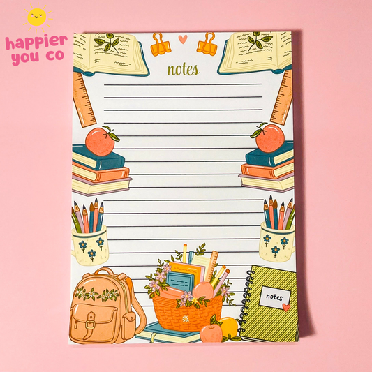A school-themed notepad with lined writing space in the center, decorated with illustrations of books, pencils, an apple, a backpack, and a basket. The notepad is shown on a solid pink background with a small 'happier you co' logo in the top-left corner.