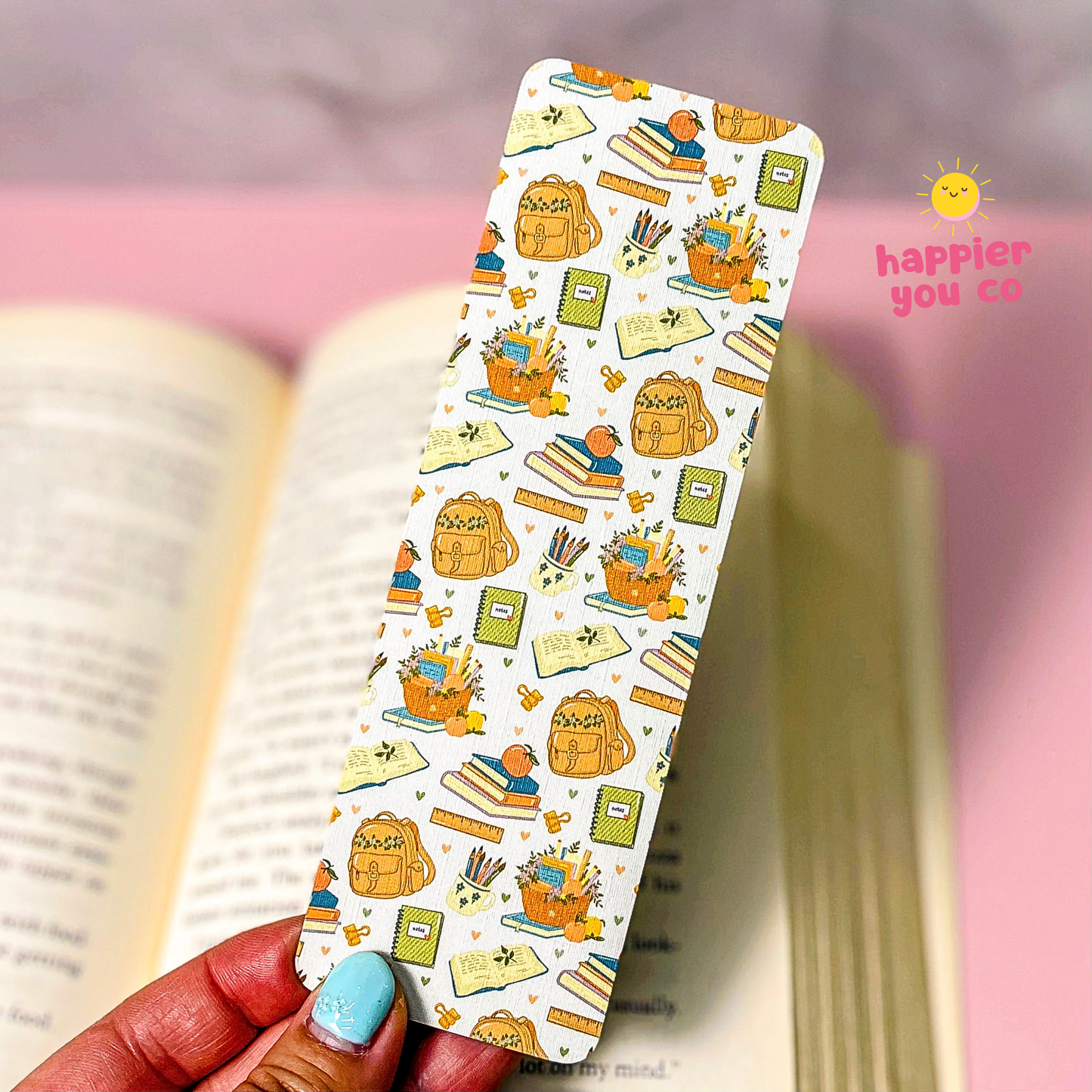 A hand with light blue nail polish holds a white bookmark decorated with a colorful school and reading-themed pattern, set against a soft-focus background of an open book and a pink surface. A 'happier you co' logo is visible in the top right corner.
