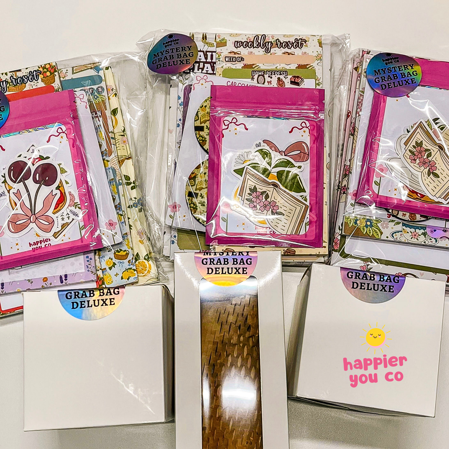 Three packaged 'Mystery Grab Bag Deluxe' stationery sets arranged on a white surface, with their white envelopes displayed in front. The packages feature colorful floral and botanical illustrations, patterned washi tapes, and decorative stickers.