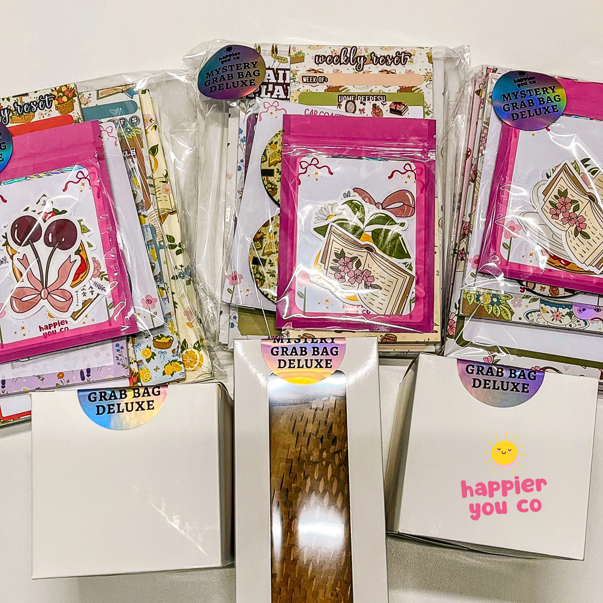 Three packaged 'Mystery Grab Bag Deluxe' stationery sets arranged on a white surface, with their white envelopes displayed in front. The packages feature colorful floral and botanical illustrations, patterned washi tapes, and decorative stickers.