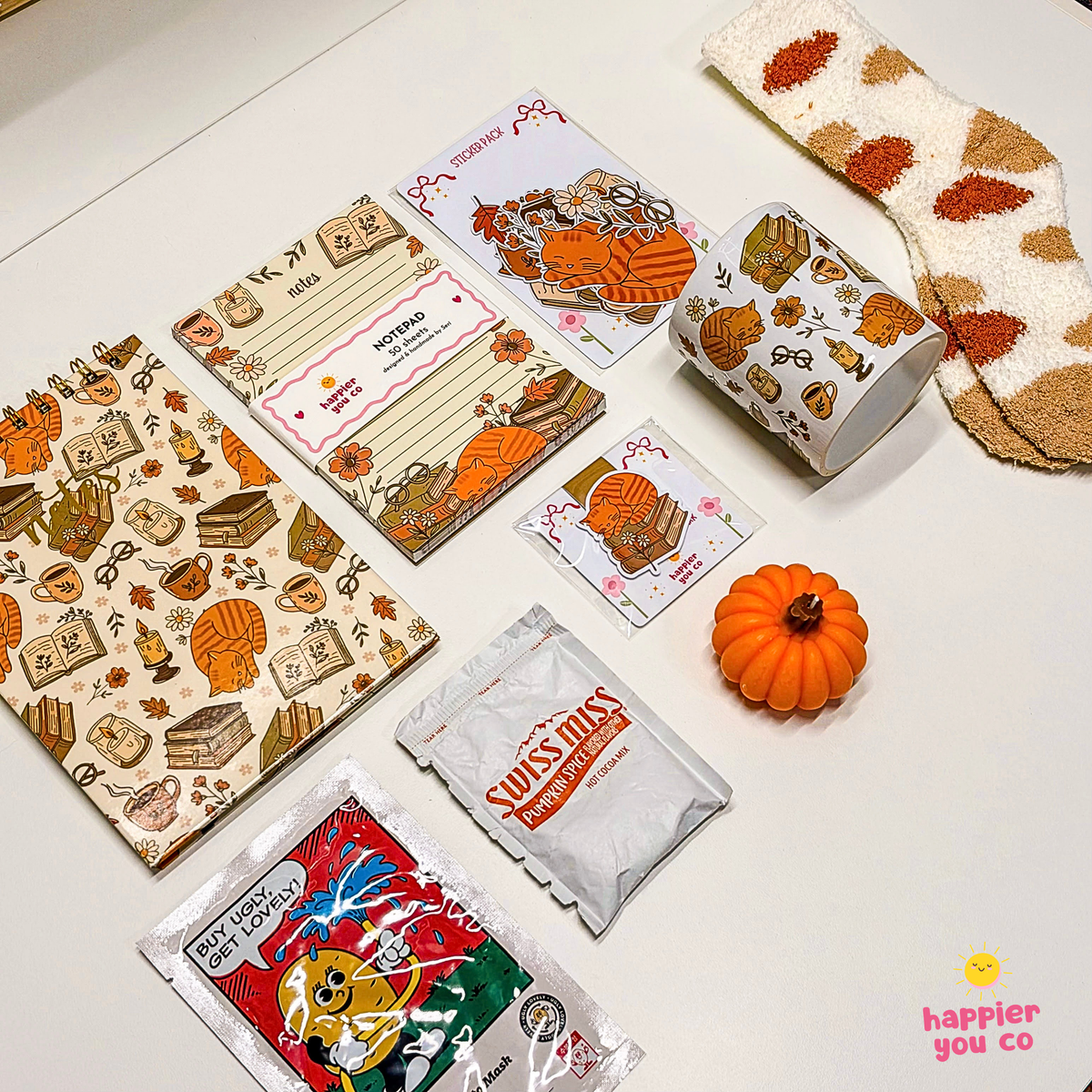 A coordinated fall-themed stationery and sock set arranged on a white surface. The set includes patterned cards, a notepad, a cup holder cover, socks, a small orange pumpkin prop, and two Swiss Miss candy packets.