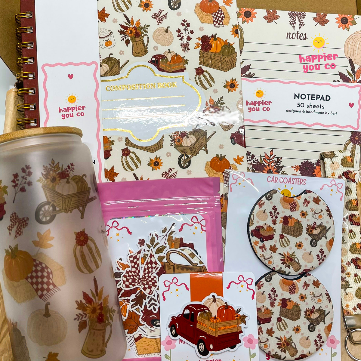 A coordinated fall-themed stationery and accessory set arranged on a light wood surface, including a patterned notepad, a lined notes pad, car coasters, sticker sheets, a cup sleeve, and a tumbler with autumn illustrations such as pumpkins, leaves, and a red truck.