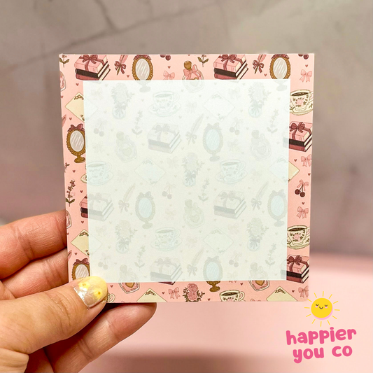 Memo pad with decorative border held by a hand, on a pink background with 'happier you co' branding.