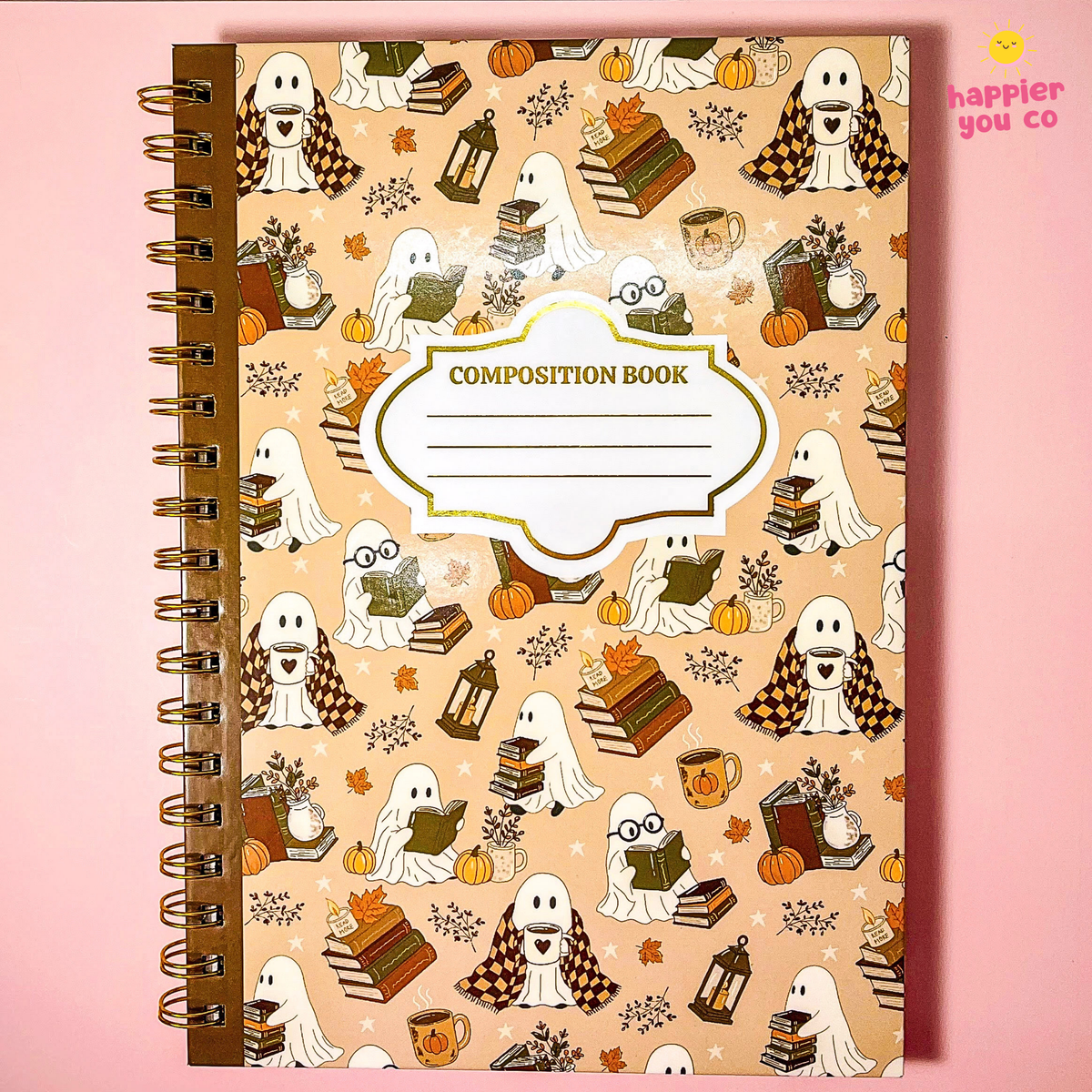 A spiral-bound composition notebook with a Halloween-themed cover featuring cartoon ghosts, books, pumpkins, and autumn leaves on a beige background, set against a solid pink backdrop.