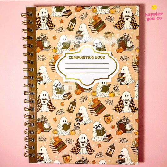 A spiral-bound composition notebook with a Halloween-themed cover featuring cartoon ghosts, books, pumpkins, and autumn leaves on a beige background, set against a solid pink backdrop.