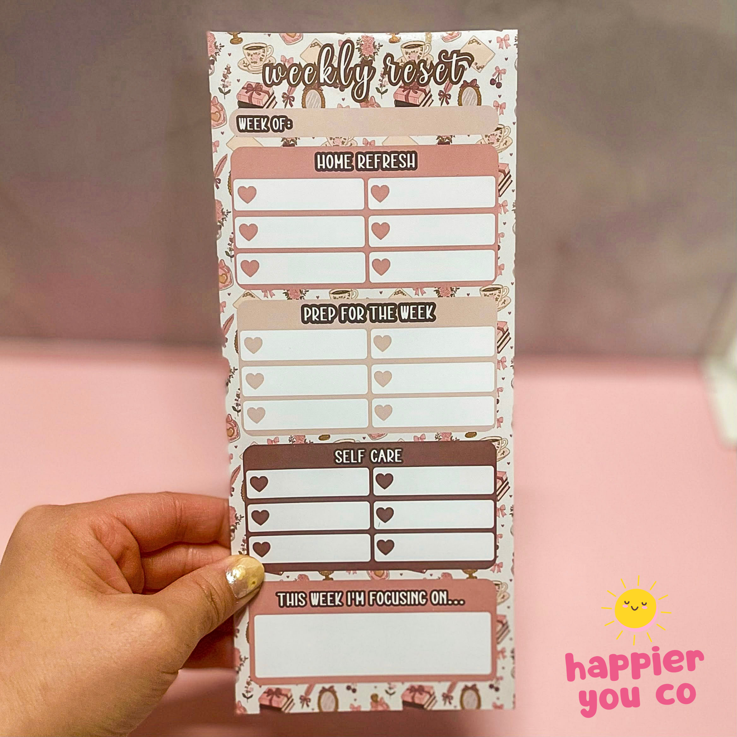 Weekly reset planner with heart designs held by a hand on a pink background, featuring 'happier you co' branding.