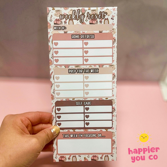 Weekly reset planner with heart designs held by a hand on a pink background, featuring 'happier you co' branding.