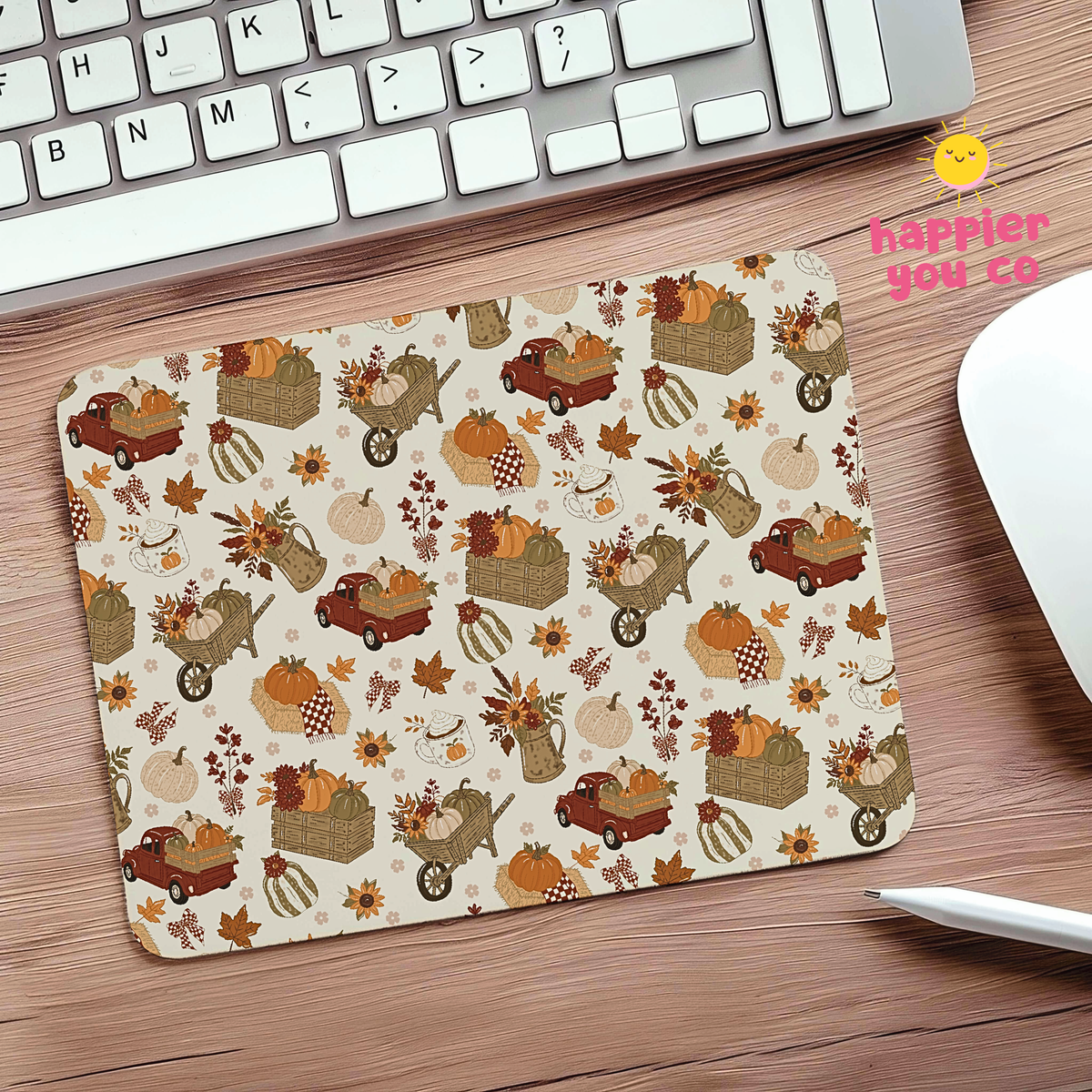 A rectangular desk mat with an autumn harvest pattern featuring pumpkins, autumn leaves, and small trucks, shown on a wooden desk next to a keyboard and mouse.