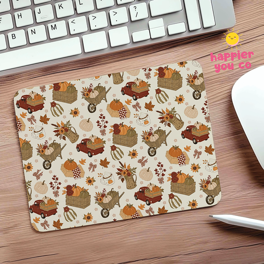 A rectangular desk mat with an autumn harvest pattern featuring pumpkins, autumn leaves, and small trucks, shown on a wooden desk next to a keyboard and mouse.