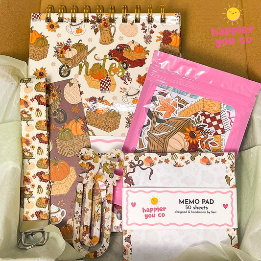 A fall-themed stationery gift set from Happier You Co inside a cardboard box, featuring a spiral notebook, patterned memo pads, a pink zip pouch, and a metal clip on white tissue paper.