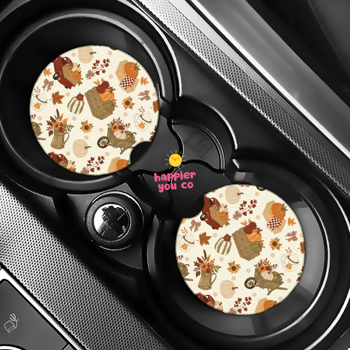 Two round car cup holder inserts with a fall-themed pattern of pumpkins, leaves, and characters on a cream background, shown installed in a black car center console. The center insert features the pink text 'happier you co'.