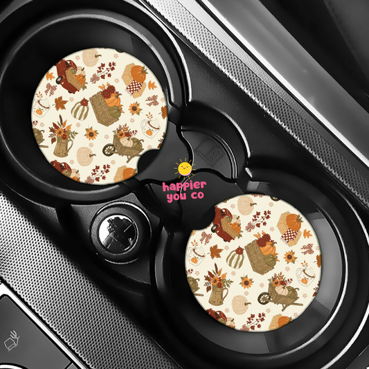 Two round car cup holder inserts with a fall-themed pattern of pumpkins, leaves, and characters on a cream background, shown installed in a black car center console. The center insert features the pink text 'happier you co'.