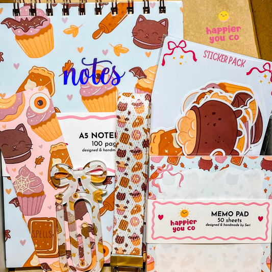 A stationery gift set featuring a spiral-bound notes notebook, a sticker pack, an A5 memo pad, and a patterned washi tape, all decorated with cute pastel illustrations of cupcakes, cats, and cookies. The products are branded with 'happier you co'.