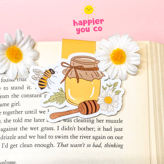 Decorative magnetic bookmark with honey jar, bee, and flowers on a book page with 'happier you' text.