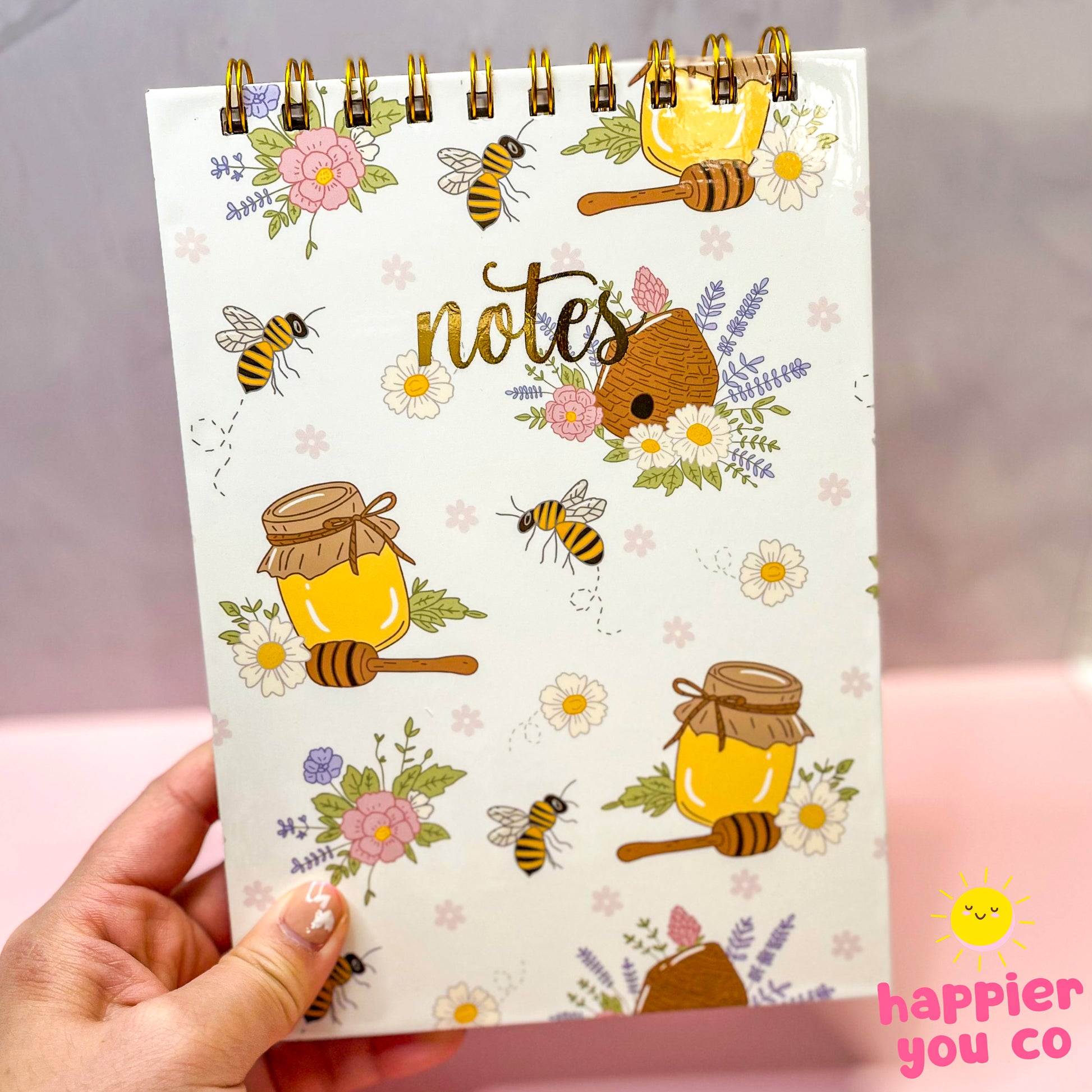 Notebook with honeybee and floral design held by a hand, featuring the brand 'happier you co'.