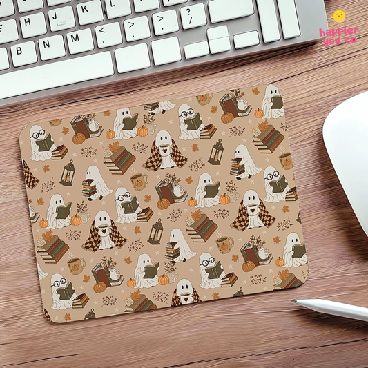 A rectangular desk mat with a repeating Halloween pattern of ghosts, pumpkins, books, and autumn leaves on a tan background, placed on a wooden desk next to a keyboard and mouse.