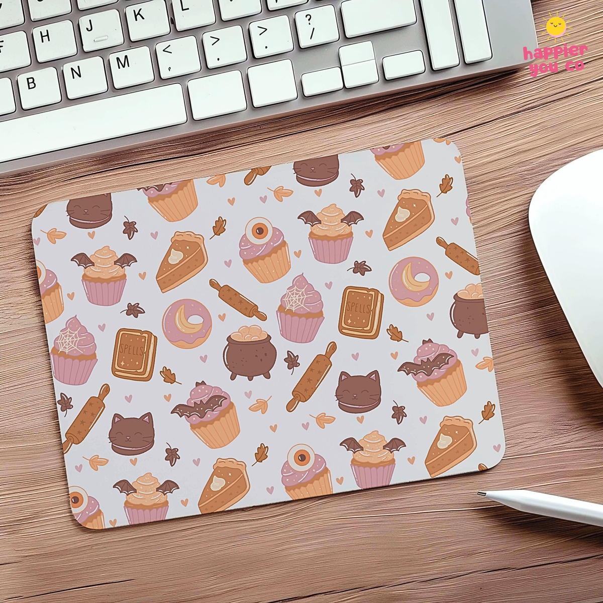 A rectangular desk mat with a cute bakery pattern of cupcakes, donuts, cakes, and rolling pins on a light background, placed on a wooden desk next to a keyboard, mouse, and stylus.