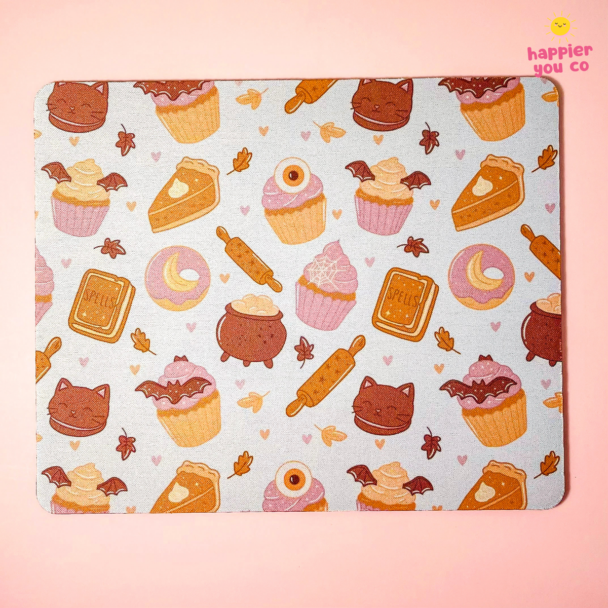 A rectangular mousepad with rounded corners featuring a cute, whimsical pattern of cupcakes, donuts, slices of cake, rolling pins, and small hearts in warm autumn colors on a pale blue-gray background. The product is displayed flat against a solid pastel pink background with a 'happier you co' logo in the top right corner.