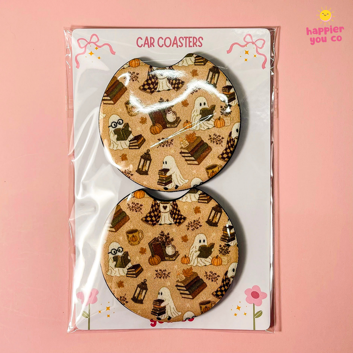 A pair of Halloween-themed car coasters in a clear plastic sleeve on a white backing card, set against a solid pink background. The coasters feature a tan background with a pattern of ghosts, pumpkins, candy corn, and autumn leaves.