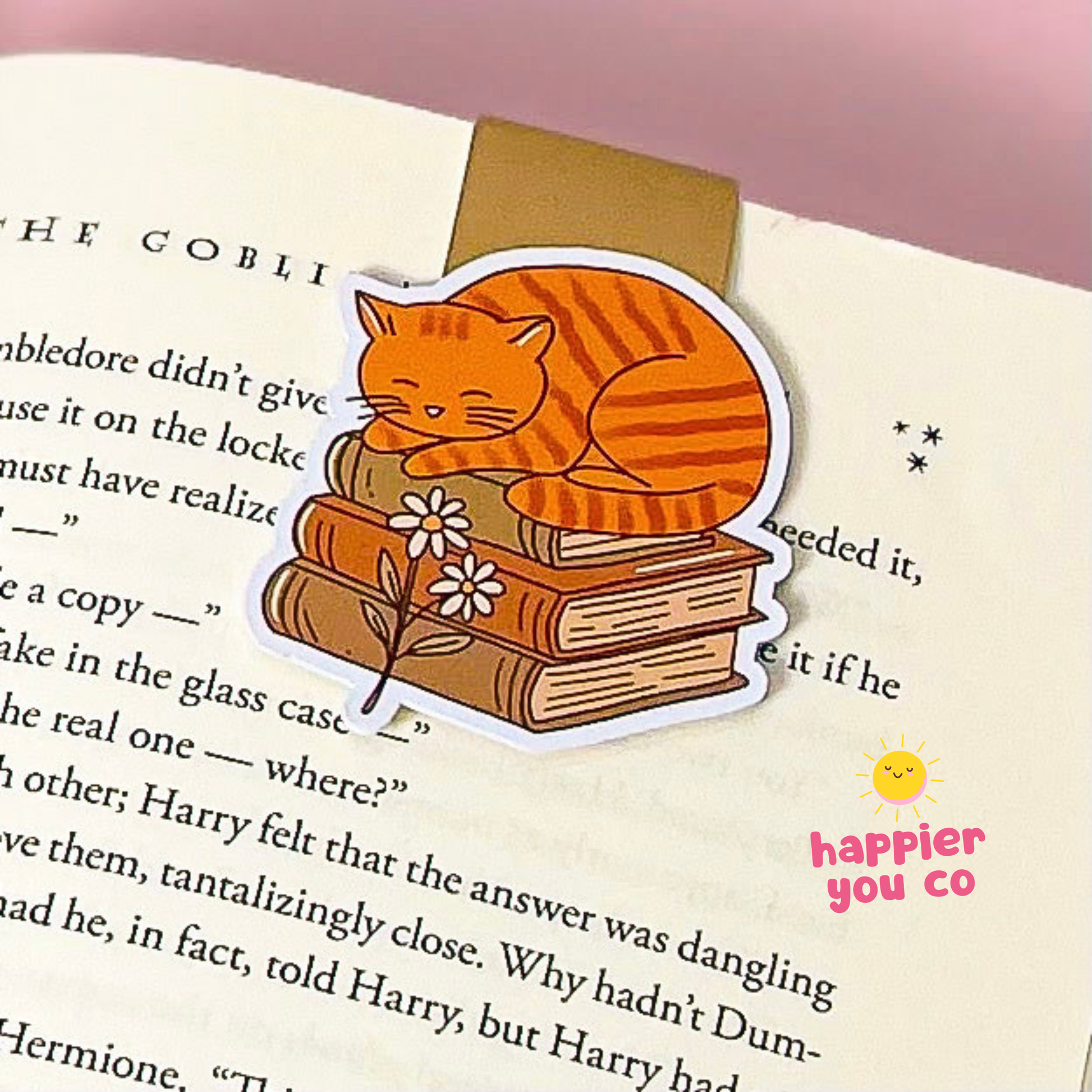 An orange tabby cat magnetic bookmark sleeping on a stack of books with a small flower, shown clipped onto an open book page. The sticker has a white border and is photographed on a pink background.