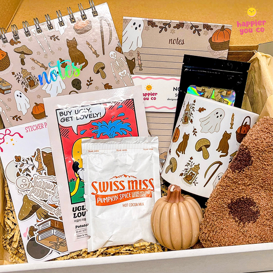 A cozy fall-themed gift box filled with stationery and treats. The box contains a spiral notebook, a notes pad, stickers, a sticker pack, a bag of 'Swiss Miss Pumpkin Spice Hot Cocoa Mix', and a brown plush toy, all arranged in paper shred.