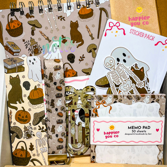 A curated stationery gift set from happier you co featuring a sticker pack, memo pad, notebook, and washi tape with a cohesive autumn skeleton and cat pattern.