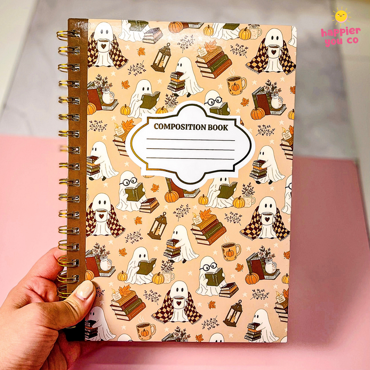 A hand holds a spiral-bound composition notebook with a Halloween-themed cover featuring cartoon ghosts, books, pumpkins, and autumn motifs on a peach background. The notebook has a brown spiral binding and a white label area that reads 'COMPOSITION BOOK'.