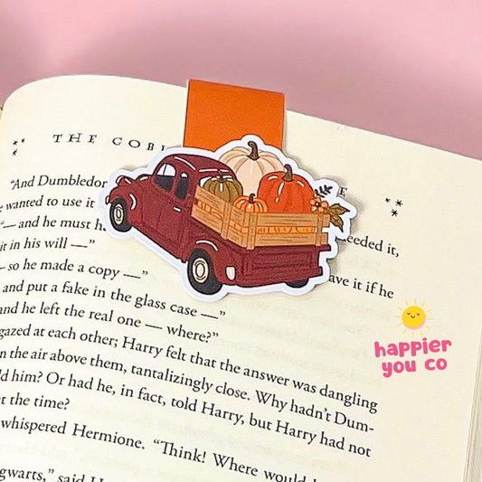 A magnetic bookmark featuring a vintage red pickup truck carrying pumpkins and a pumpkin on its bed, placed on an open book with a pink background. A yellow 'happier you co' logo is visible in the bottom right corner.