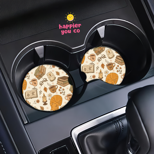 Two round car cup holder coasters with a cozy autumn coffee-themed pattern, shown installed in a black car center console. The pattern features illustrations of a pumpkin, mug, book, and coffee cups on a cream background.