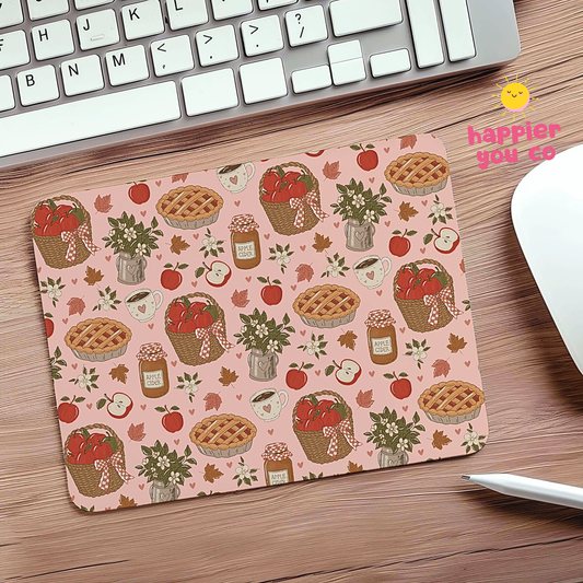 A rectangular desk mat with a pink background and a repeating autumn apple-themed pattern, shown on a wooden desk next to a keyboard, mouse, and stylus. The pattern includes apple pies, baskets of apples, jars, leaves, and small floral arrangements.