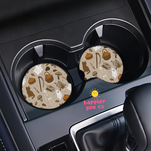 Two round car cup holder coasters with a Halloween-themed pattern of ghosts, pumpkins, cats, and bats on a beige background, shown installed in a car's cup holders.